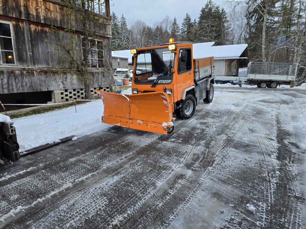 Ein orangenes Fahrzeug, welches den Schnee vor einem Haus räumt.